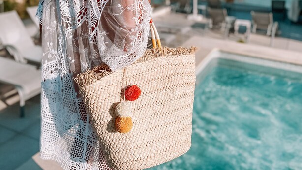 A person in a white crochet dress and straw hat, holding a straw bag with a red fruit, standing by a pool with a view of the sea and buildings.