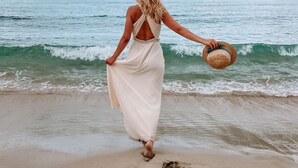 A woman in a white dress walking on a beach, holding a straw hat.