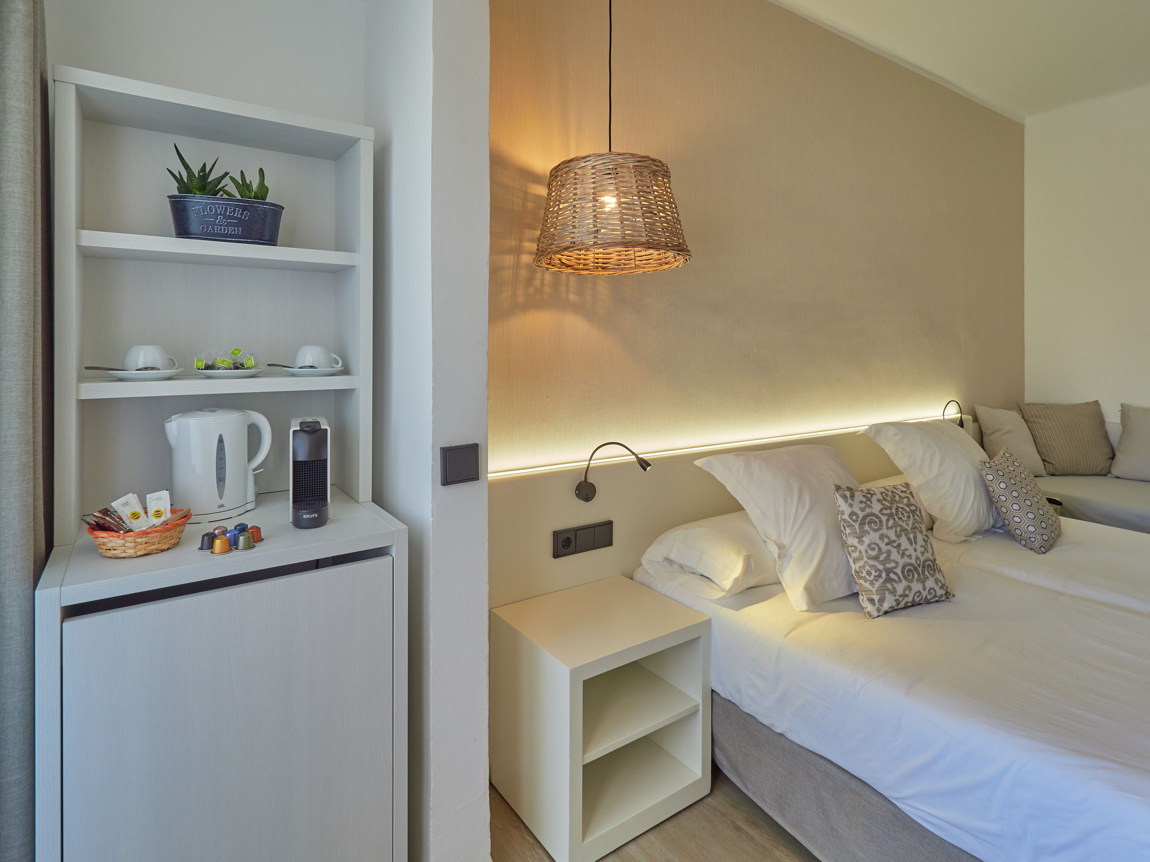 A modern hotel room with a bed, bedside tables, a shelf with a potted plant, a kettle, and a basket of toiletries.