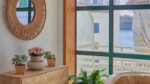 A room with a large window, a wooden cabinet with potted plants, and a woven mirror on the wall.