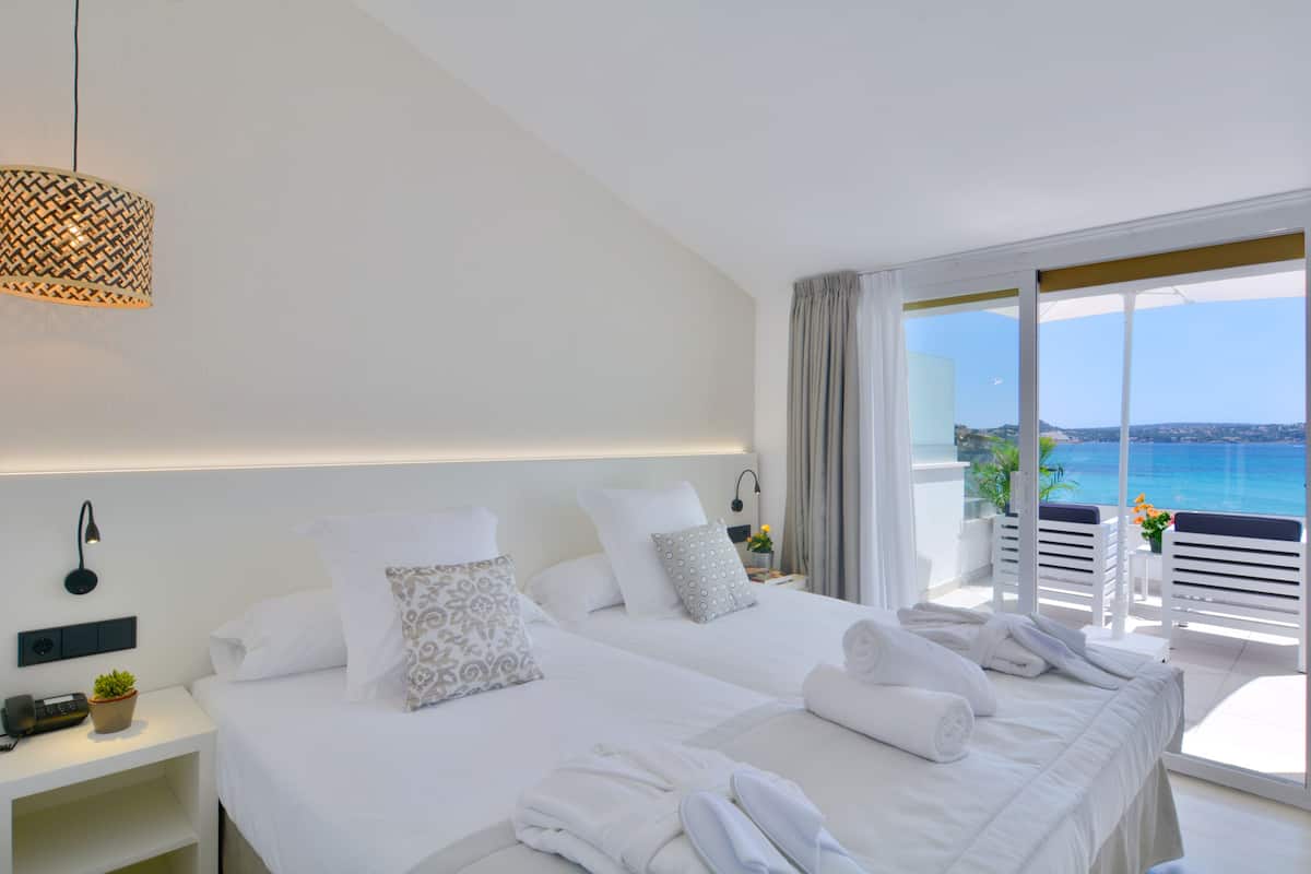 A hotel room with two beds, a bedside table with a radio and a plant, and a view of the sea through large windows.