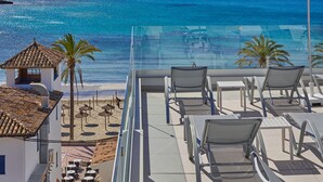 A balcony with a clear view of the sea, a palm tree, and a small building with a tiled roof.