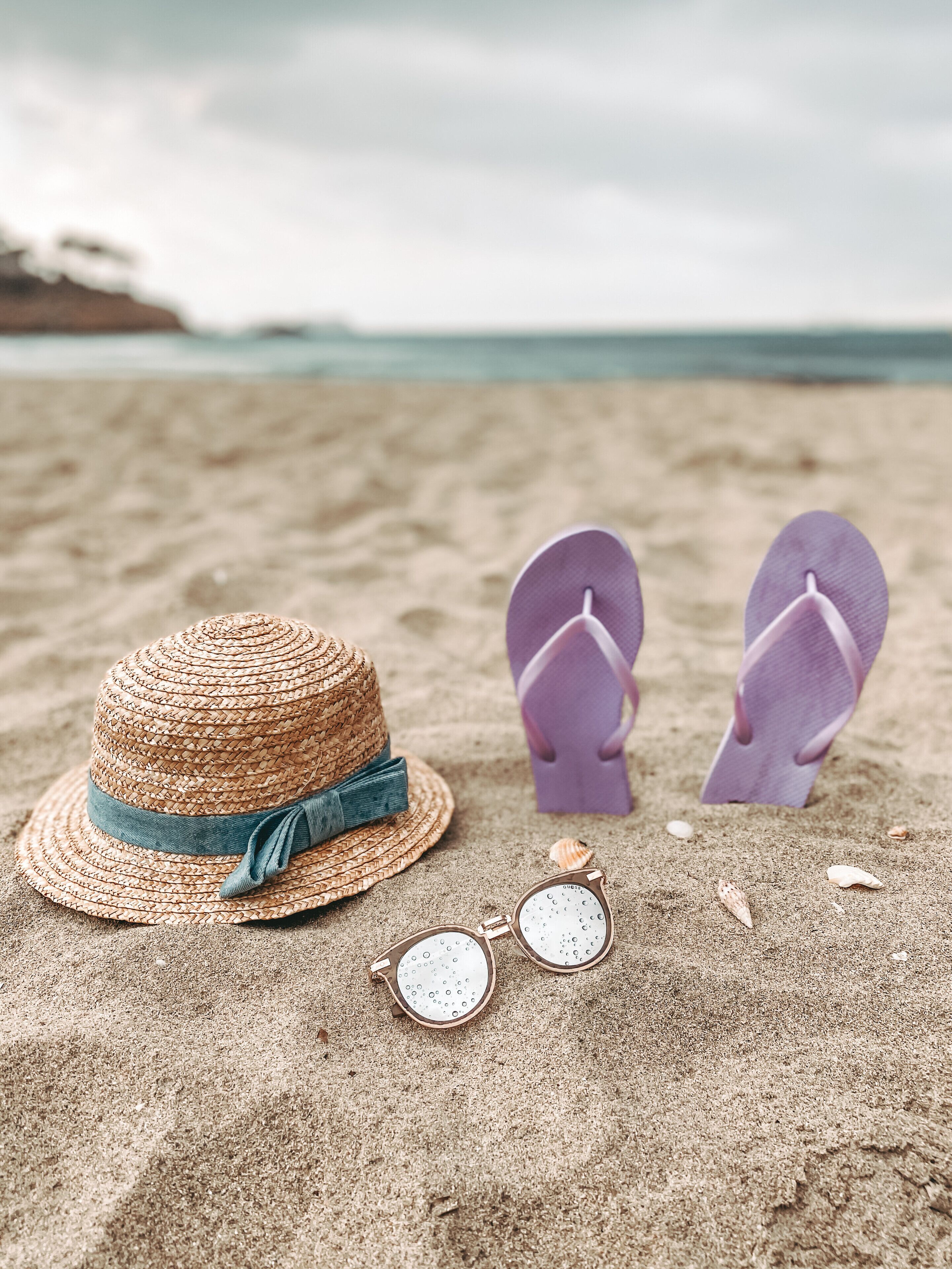 A straw hat, flip-flops, and sunglasses on a sandy beach.