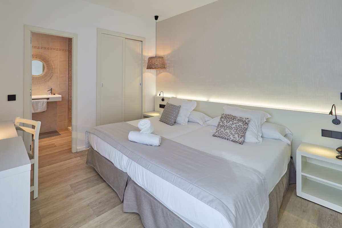 A modern bedroom with a large bed, bedside tables, a nightstand, a hanging light, a mirror, and a bathroom with a sink and towel rack.