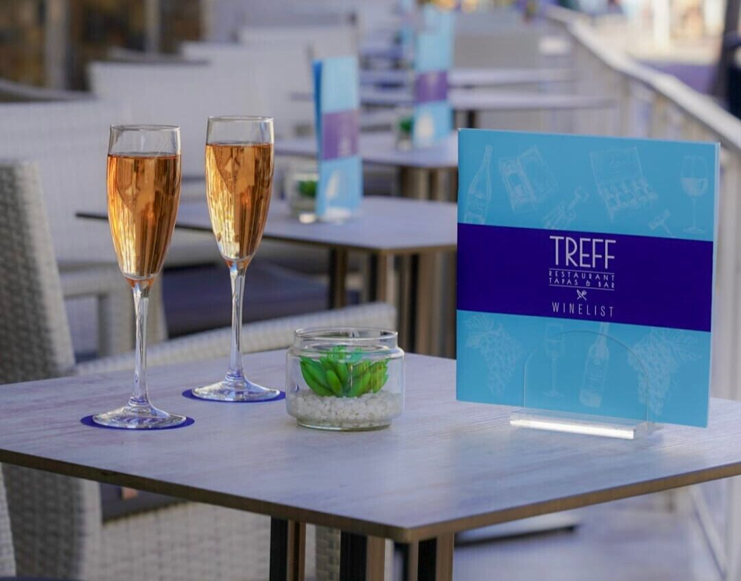 A table with two champagne flutes, a small glass jar with green plants, and a menu stand with the text "TREFF FESTAURANT TATAS & SAL."