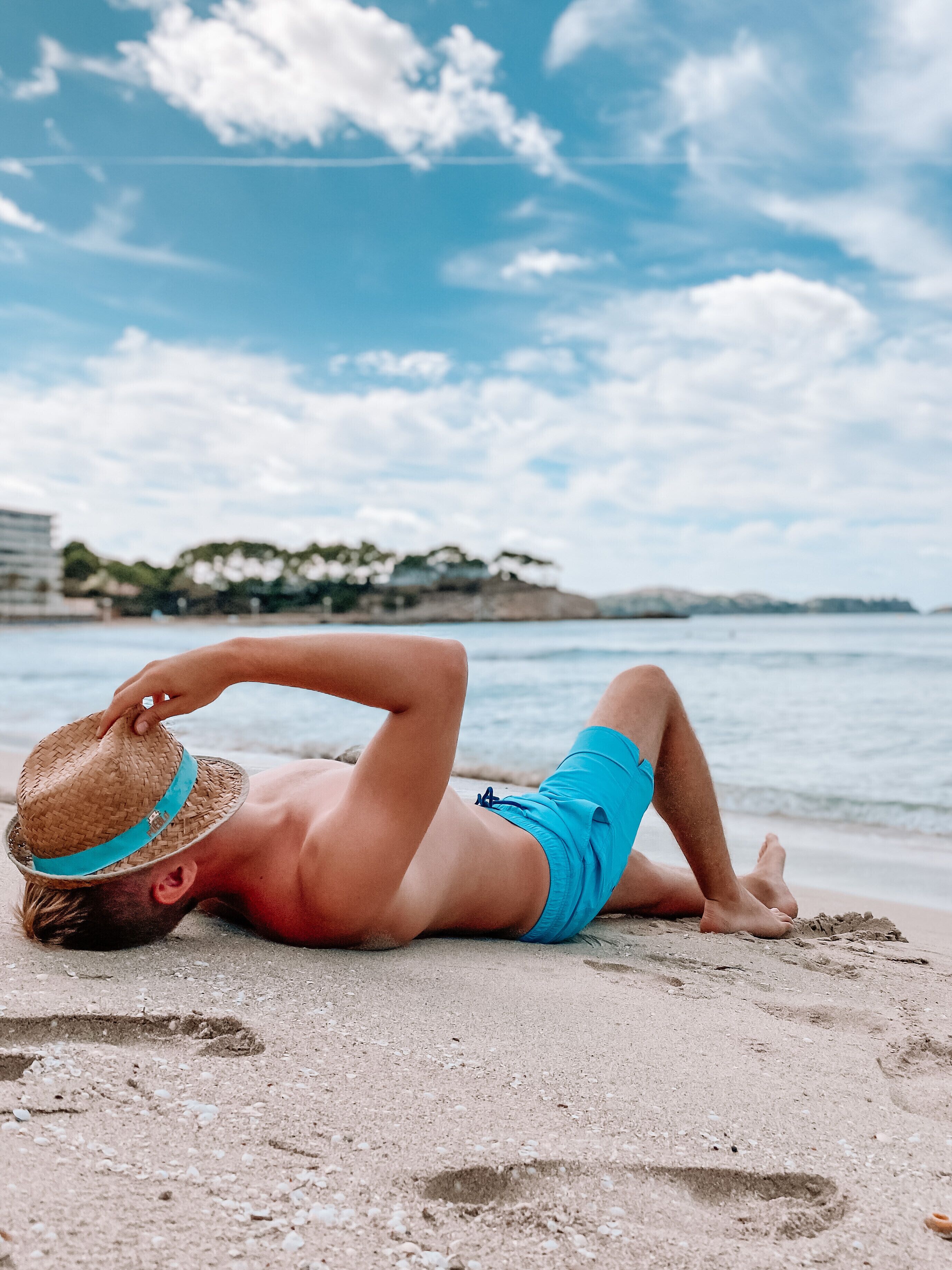 A person lying on a sandy beach with a hat on their head, wearing blue swim trunks.