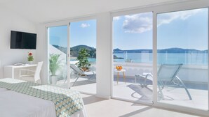 A modern hotel room with a large bed, a desk, a TV, and a balcony with a view of the sea.