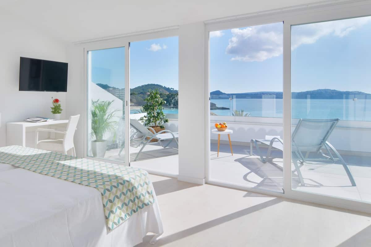 A modern hotel room with a large bed, a desk, a TV, and a balcony with a view of the sea.