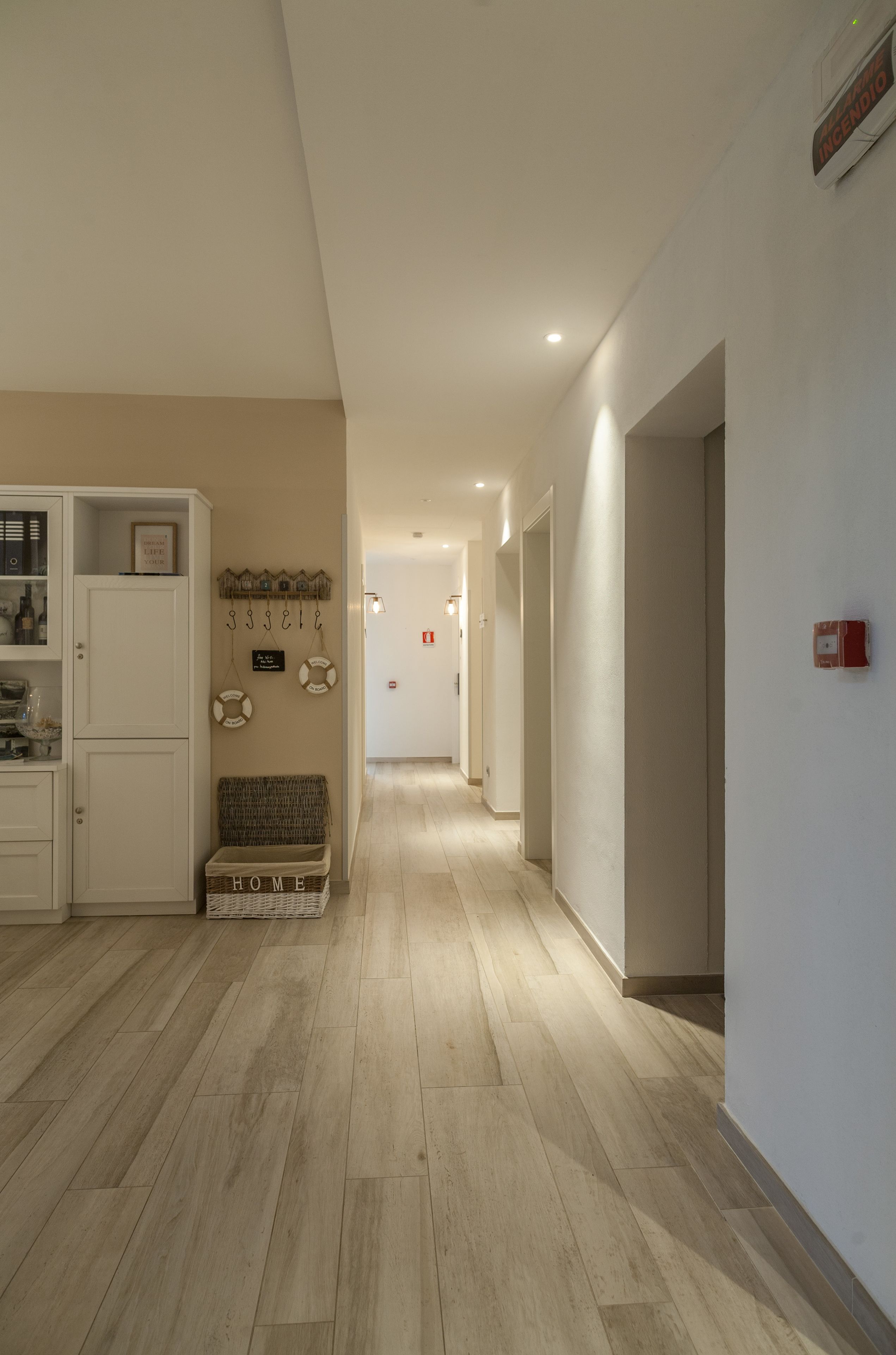 Een hal met een houten vloer, een wit kastje, een rieten bankje en een bord aan de muur.