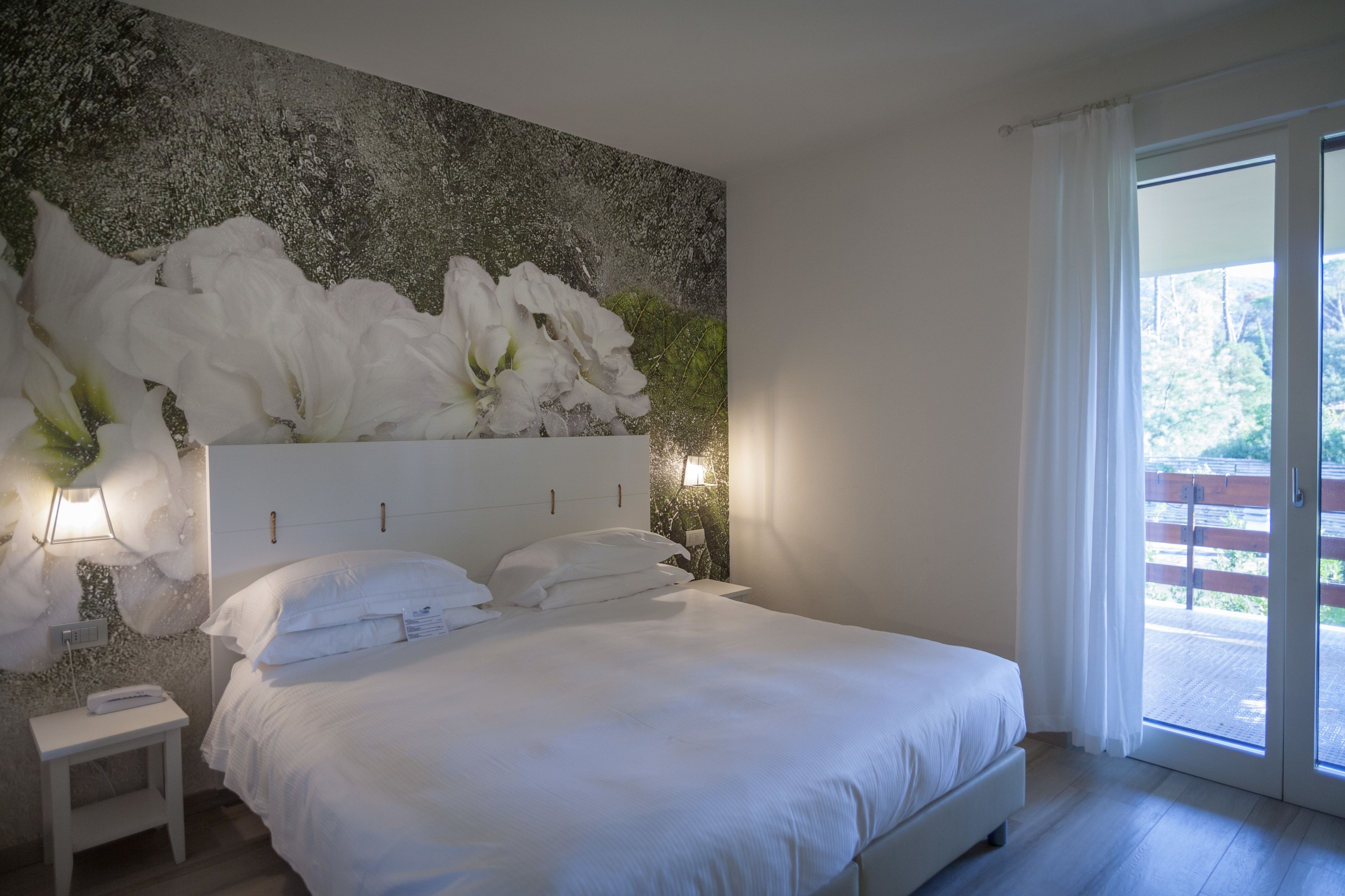 A bedroom with a large bed, a nightstand, a wall-mounted light, and a view of the outdoors through a sliding glass door.