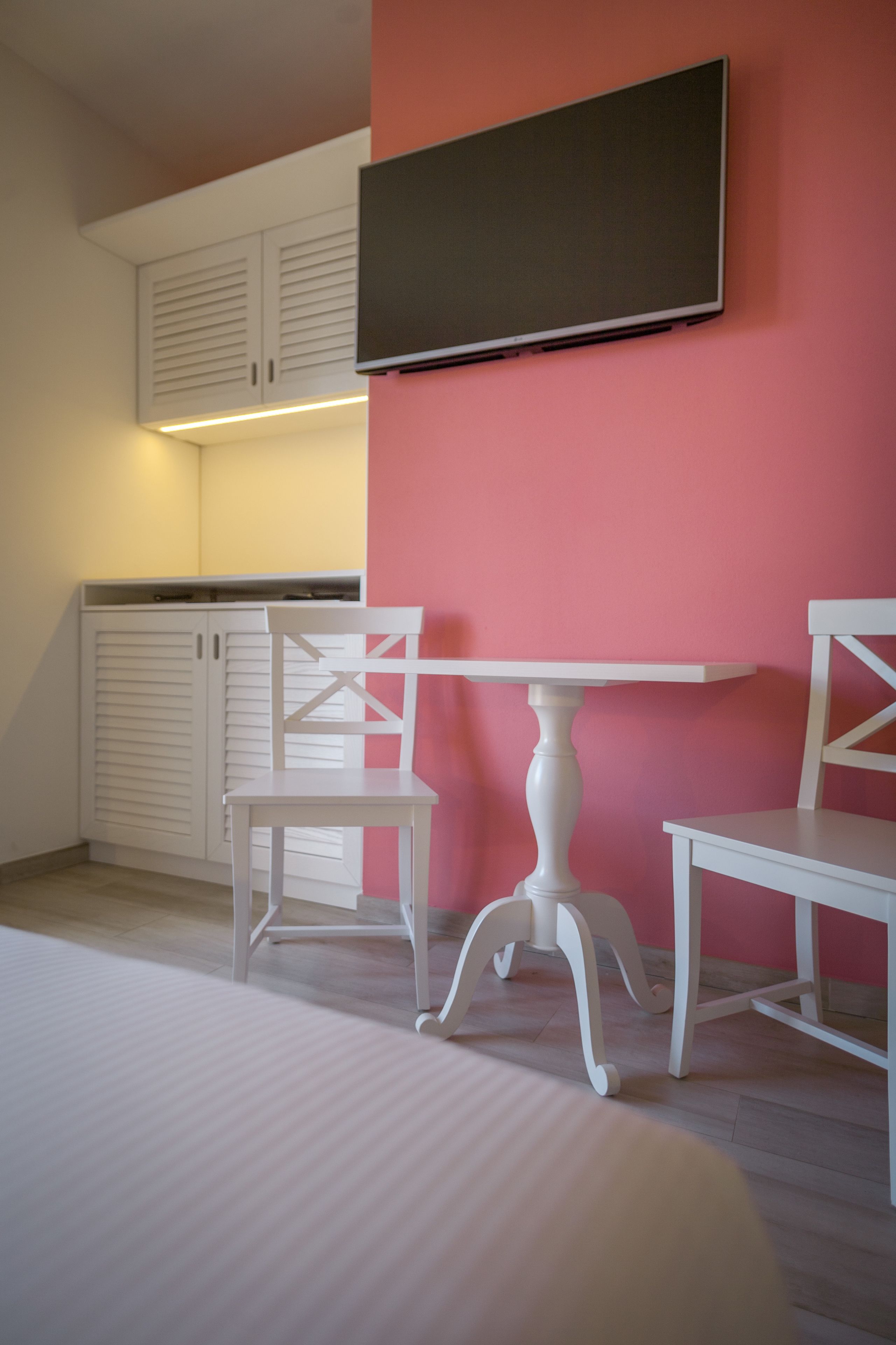 A room with a white table and two chairs, a flat-screen TV mounted on the wall, and white cabinets with a slatted design.