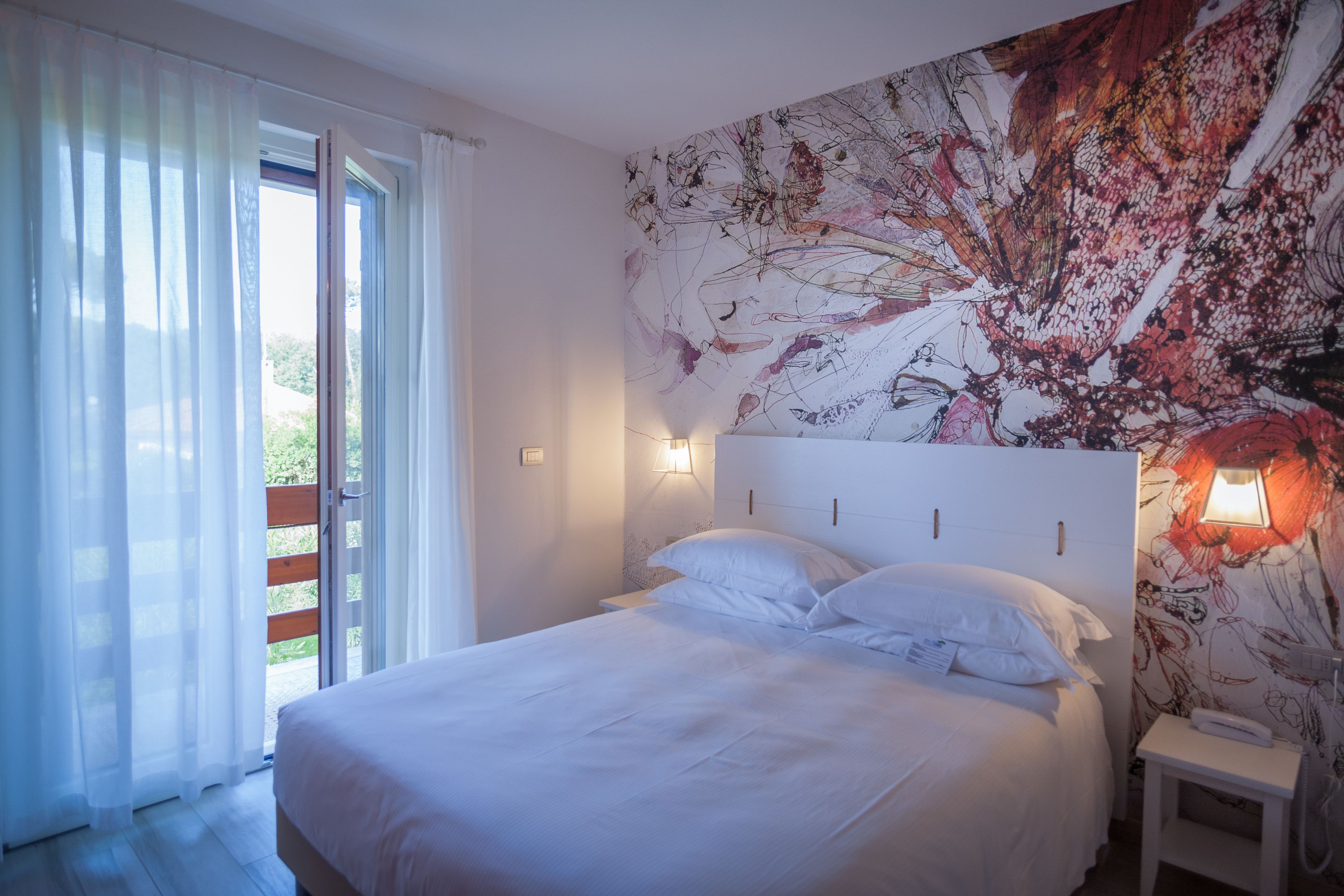 A bedroom with a large bed, a nightstand, a wall-mounted lamp, and a view through a sliding door to an outdoor area.