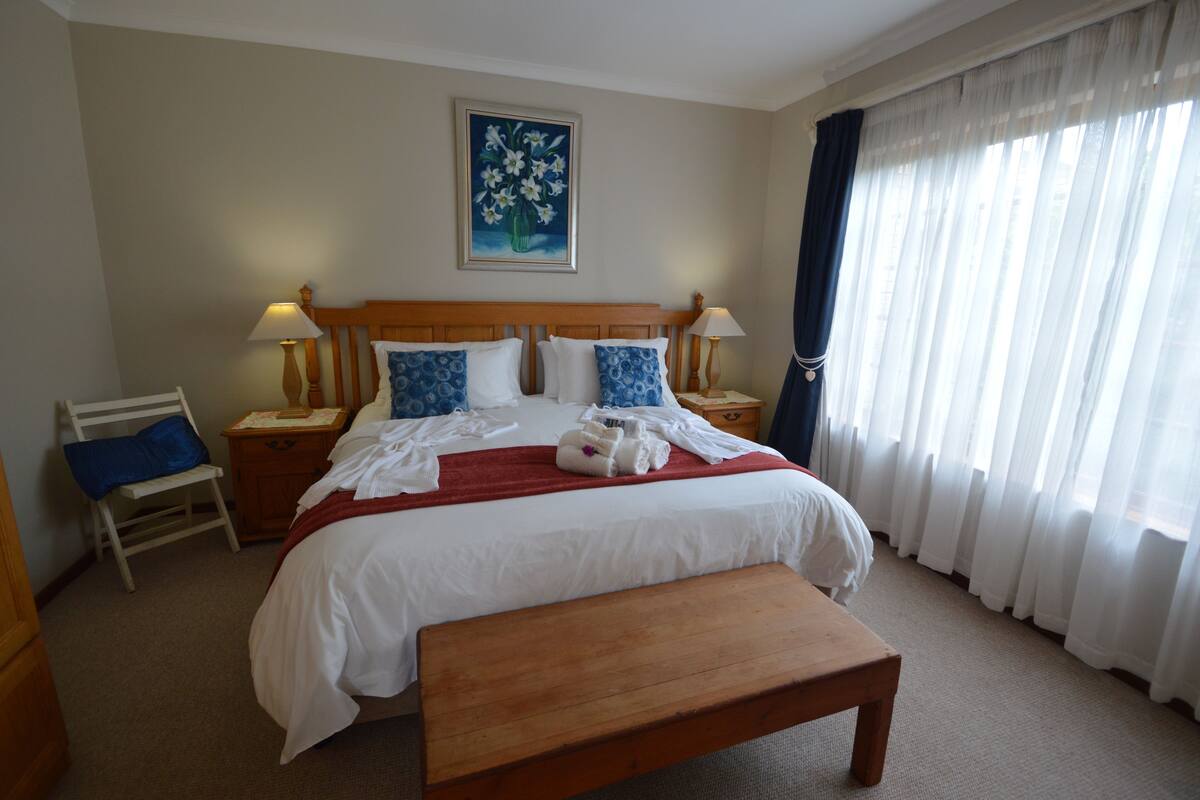A hotel room with a large bed, a wooden bench, a nightstand with a lamp, a chair, and a painting on the wall.