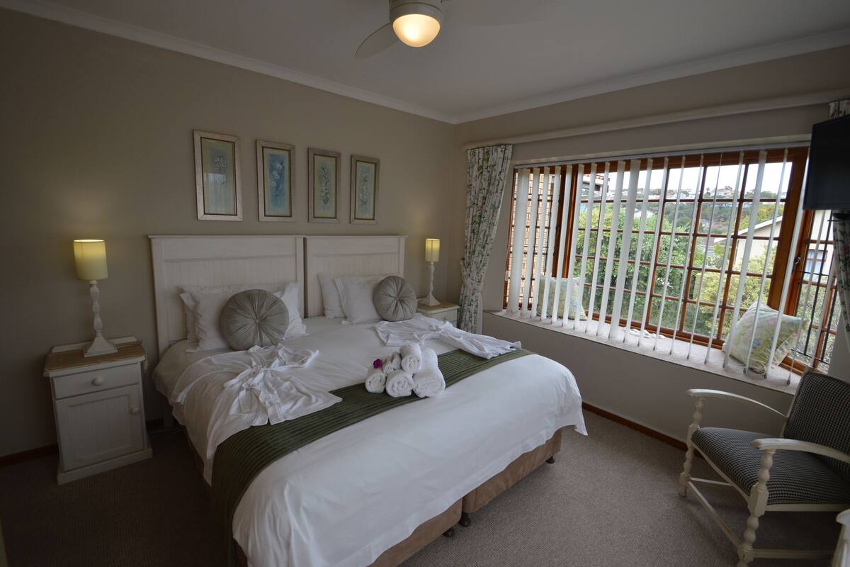 A neatly made bed with pillows and a blanket, a bedside table with a lamp, a chair, and a window with blinds.