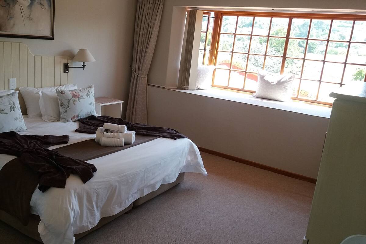 A hotel room with a large bed, a nightstand, a lamp, and a window with a view of greenery.