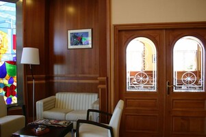 A hotel lobby with a wooden paneled wall, a stained glass window, a sofa, a chair, and a door with a circular window.