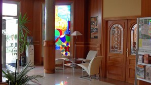 A hotel lobby with a large stained glass window, a reception desk, a potted plant, and a seating area with chairs. - Hotel Camino Real (Valdefresno)