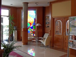 A hotel lobby with a large stained glass window, a reception desk, a potted plant, and a seating area with chairs.
