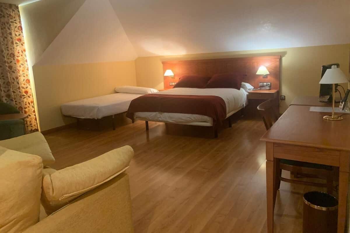 A hotel room with two single beds, a wooden table, a chair, and a small desk with a lamp.