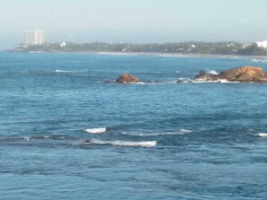 A coastal scene with clear blue water, rocky outcrops, and a distant building. - JES Guest House (Majorda)