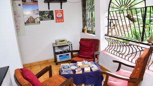 A room with a balcony, a table with books, a chair, a small shelf with decorative items, and a wall with various posters and a sign.