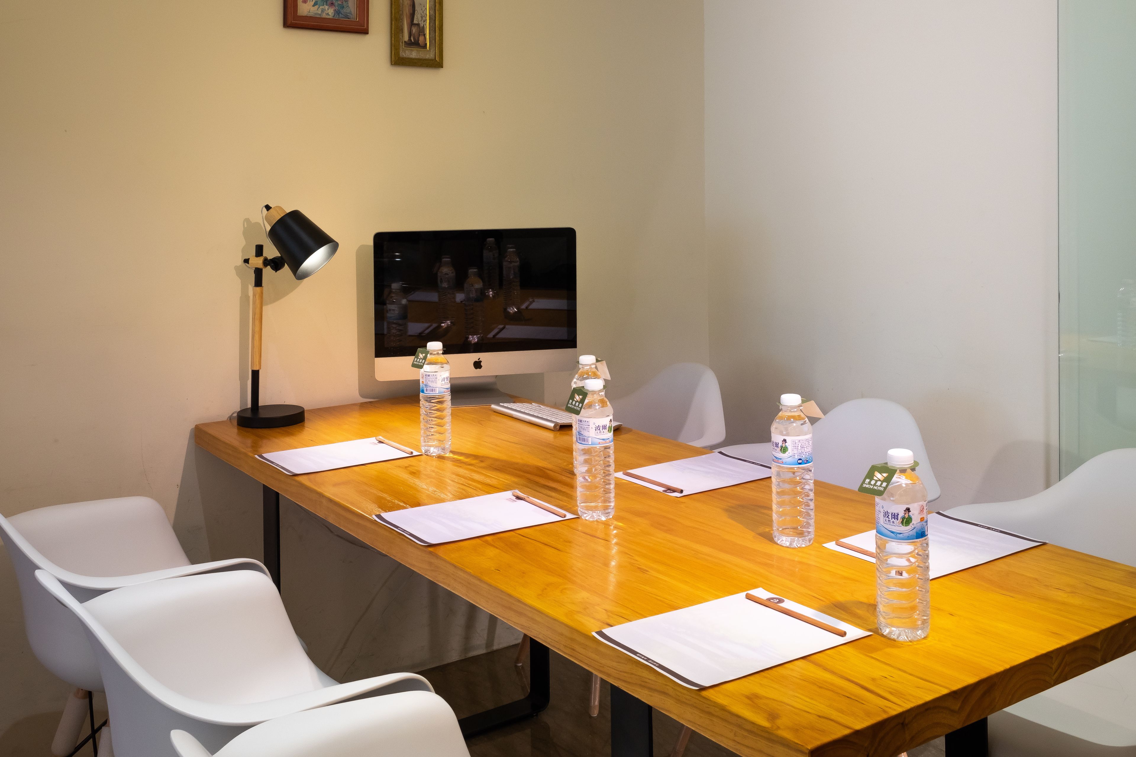A conference room with a wooden table, white chairs, a desk lamp, a computer monitor, and water bottles.