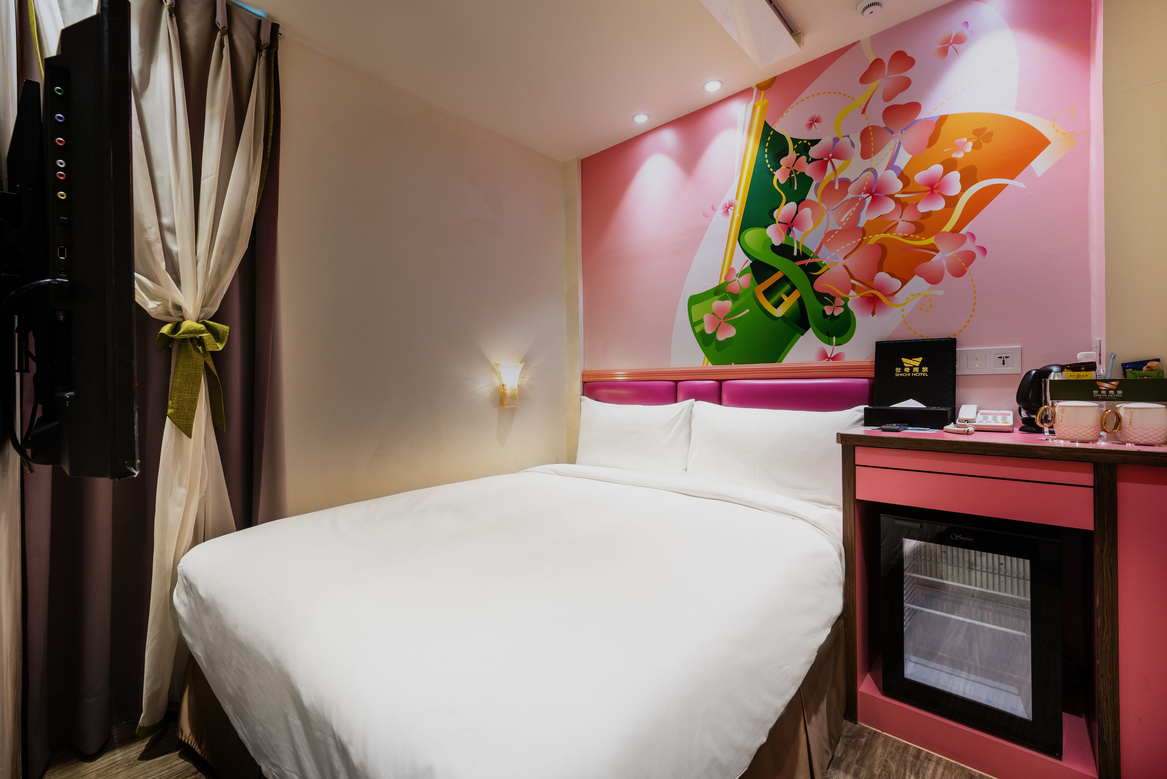 A hotel room with a bed, a desk with a computer, and a wall with a floral mural.