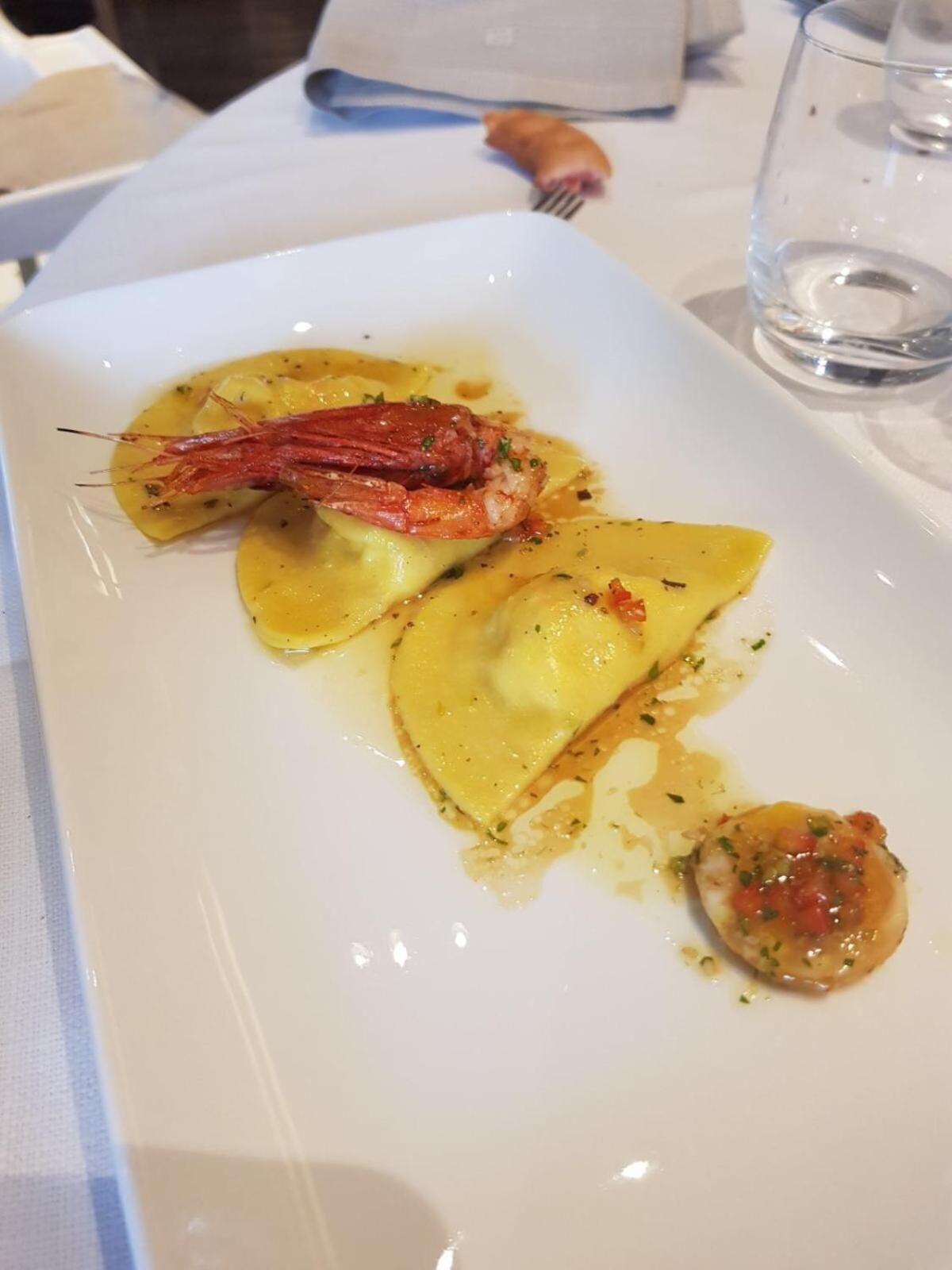 Une assiette rectangulaire blanche contenant des raviolis, une queue de homard et une petite sauce à base de tomates.