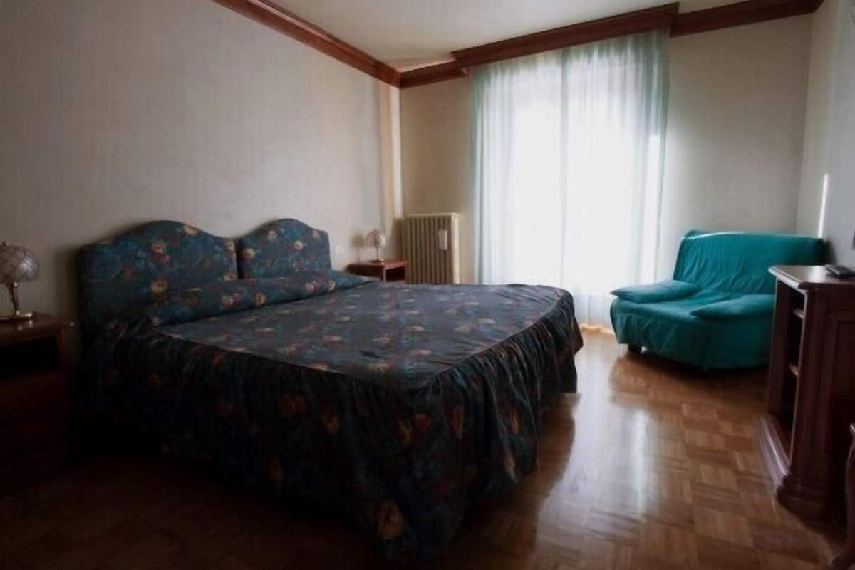 Un dormitorio con una cama grande, una silla, un radiador y una ventana con cortinas.