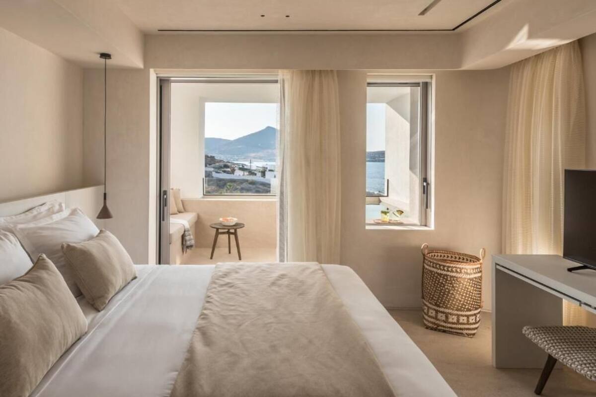 Habitación de hotel con cama, escritorio, televisión, ventana con vista al mar y a las montañas, y una pequeña mesa con una silla.