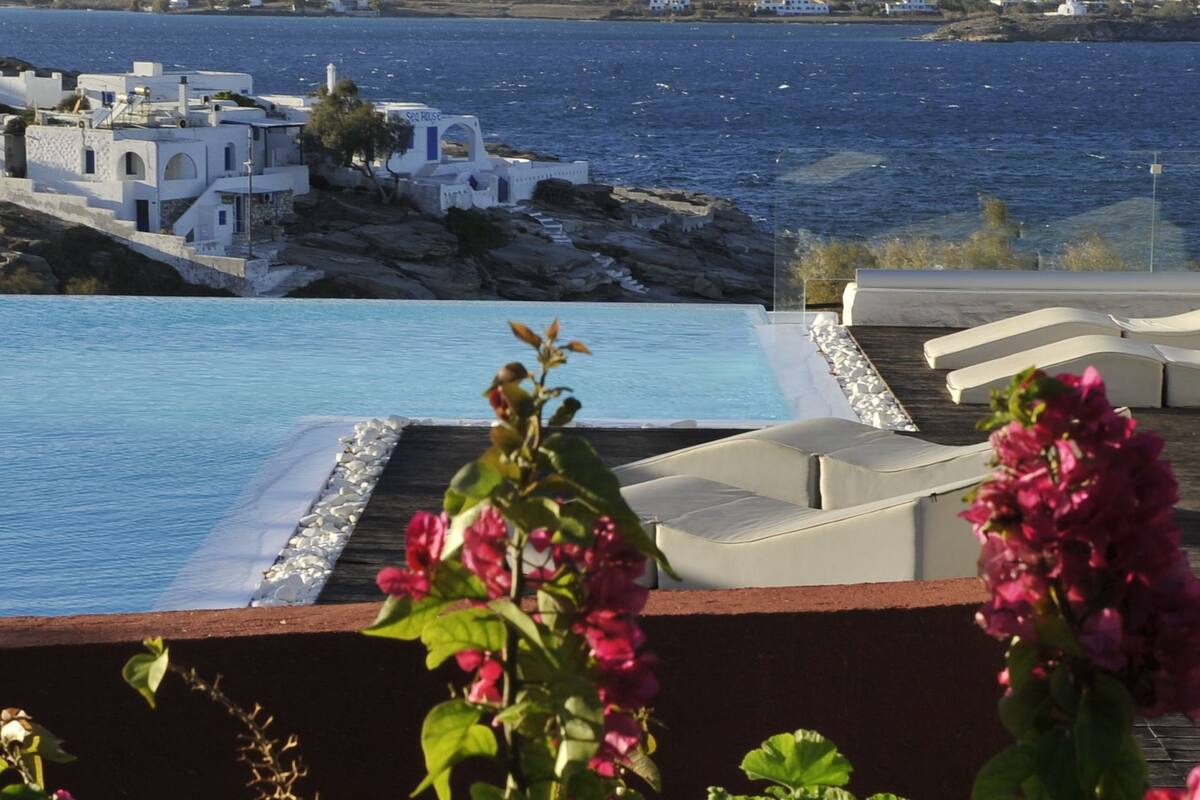Zona de piscina con sillas reclinables blancas y flores rosadas, con vistas a una costa rocosa y edificios blancos.