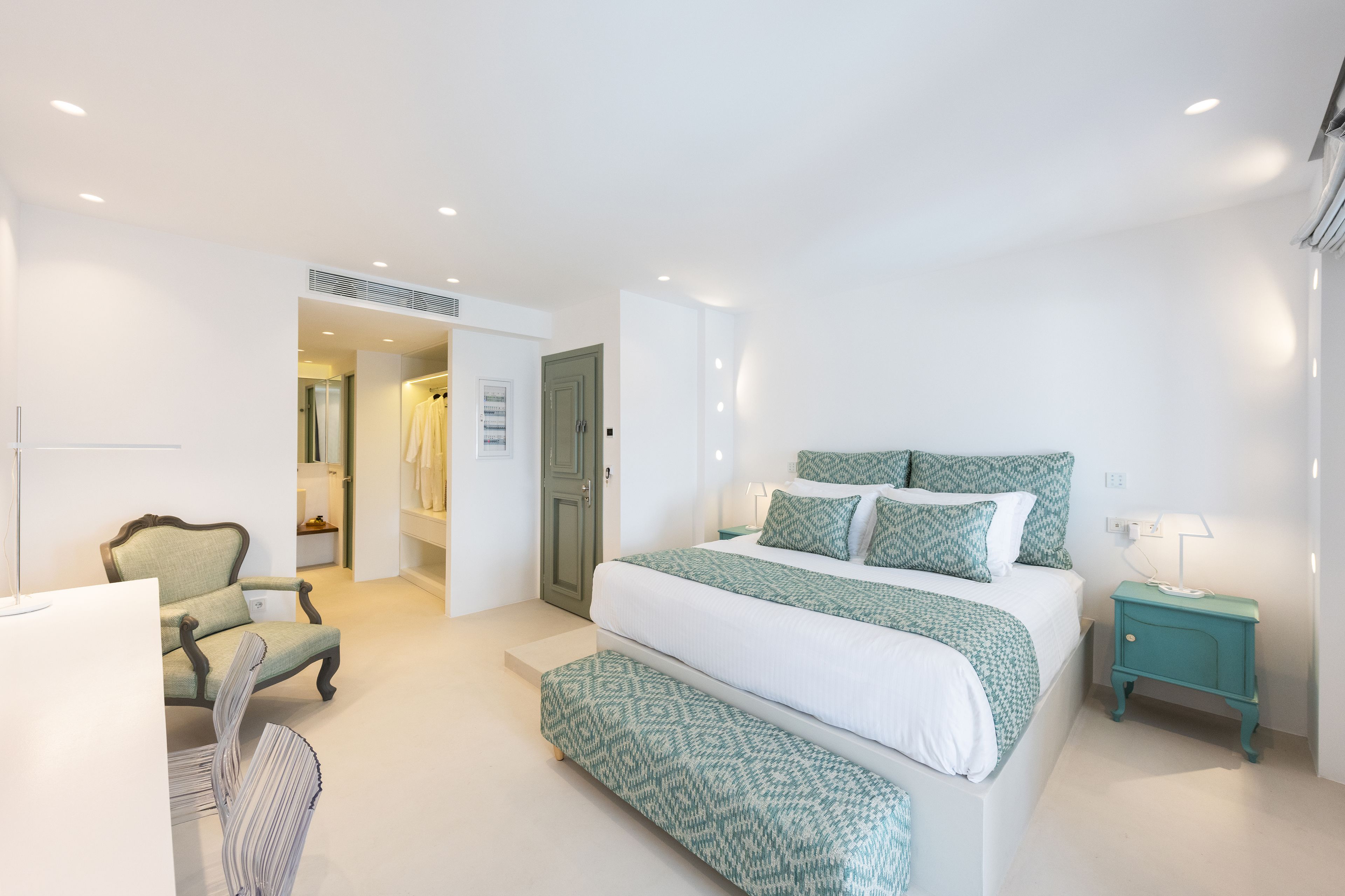 A bedroom with a large bed, a chair, a bedside table, and a door leading to another room.