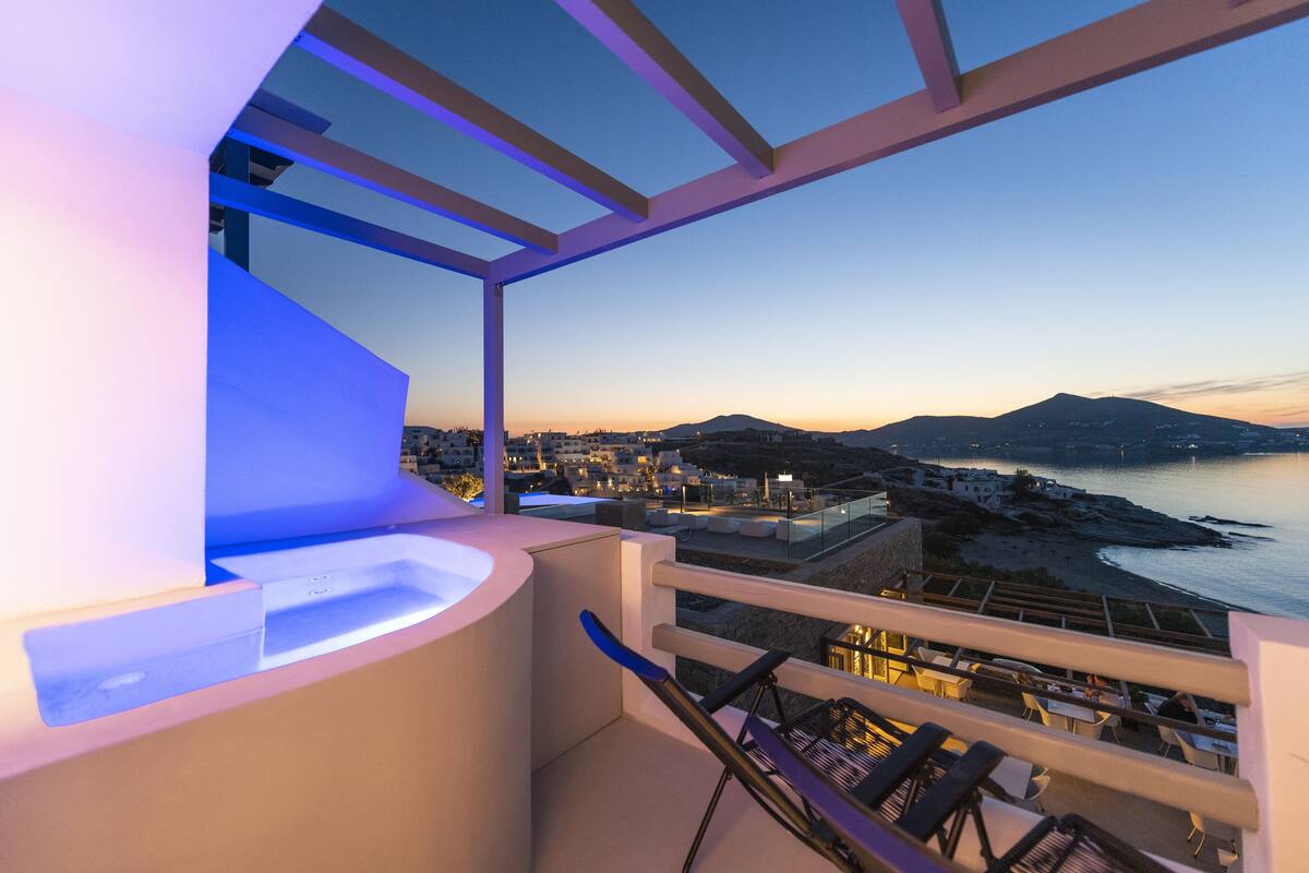 Un balcón con vista a un pueblo costero al anochecer, que presenta un diseño arquitectónico moderno con un banco curvo y una pequeña mesa.