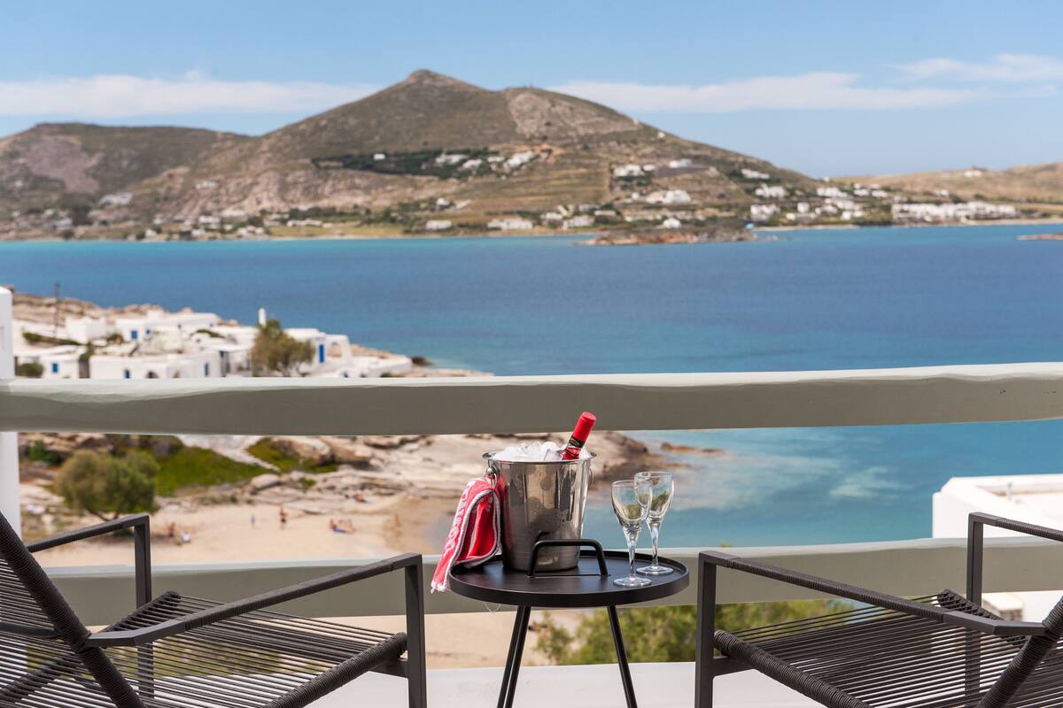 Un balcón con una mesa, una botella de champán y dos copas. Se ve una playa y una montaña.