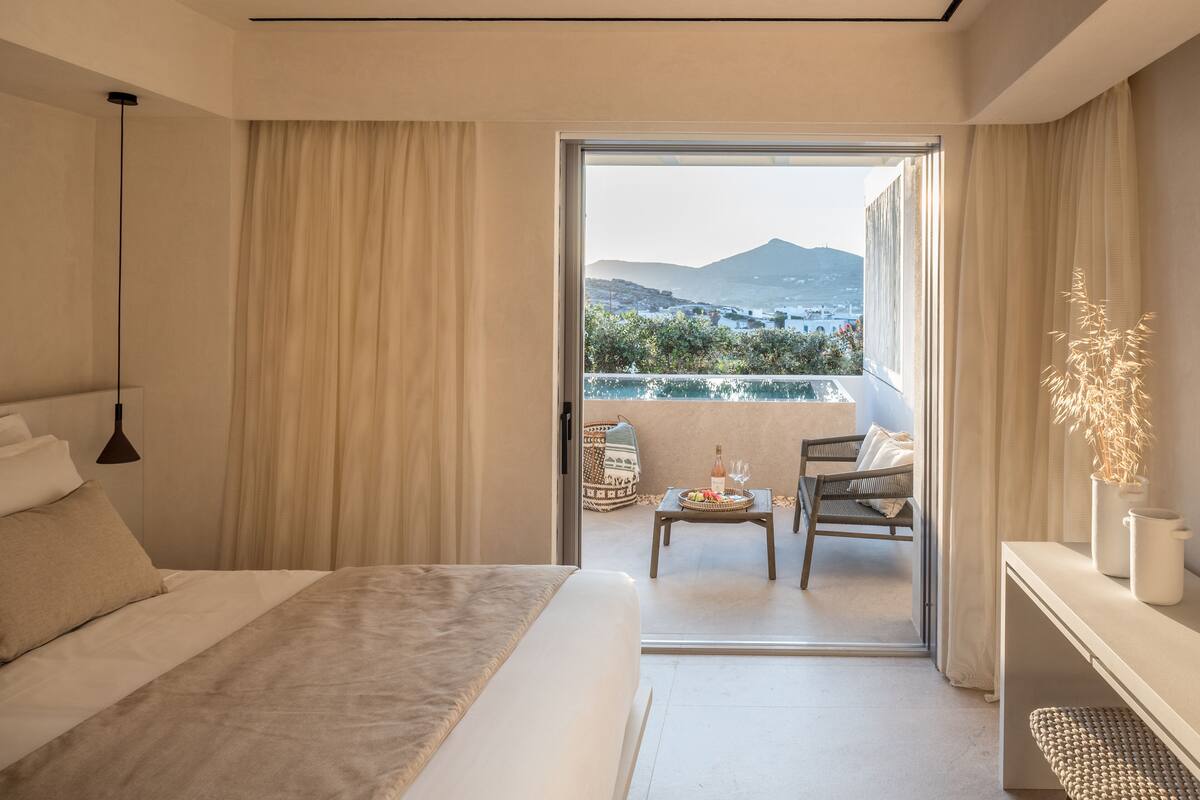 Habitación de hotel con cama, una mesita con una botella y vasos, una silla y vistas a la montaña y la piscina exterior.