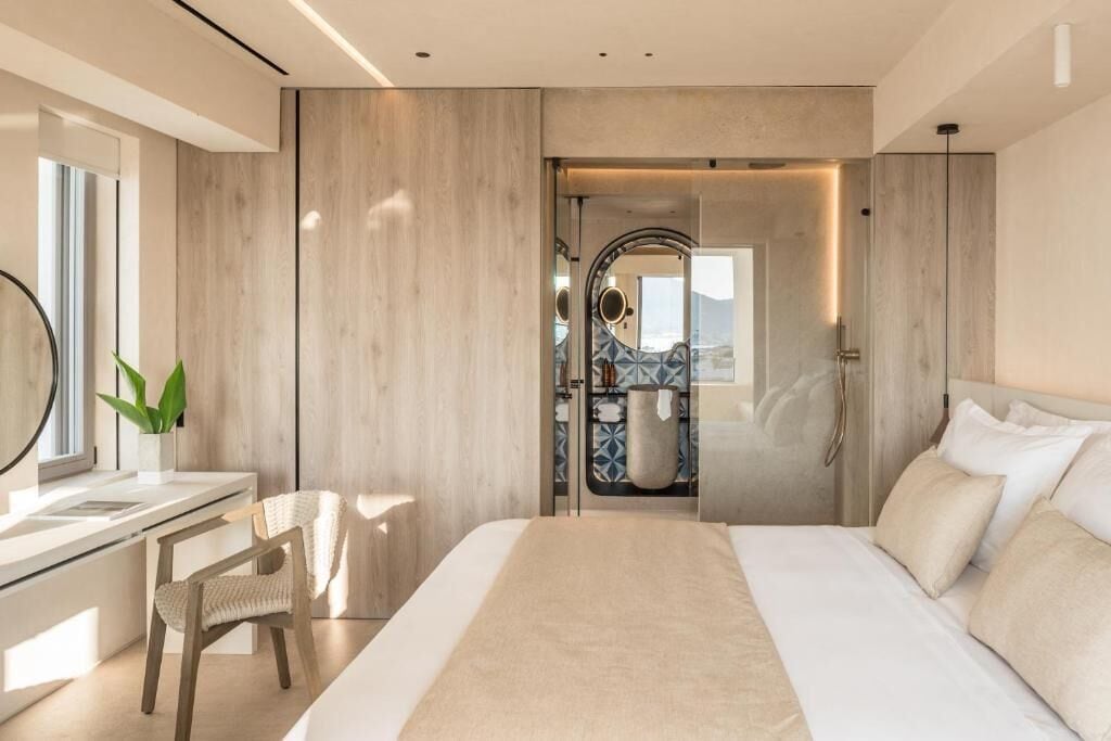 A modern hotel room with a large bed, a desk with a chair, a bathroom with a mirror, and a view of the outdoors.