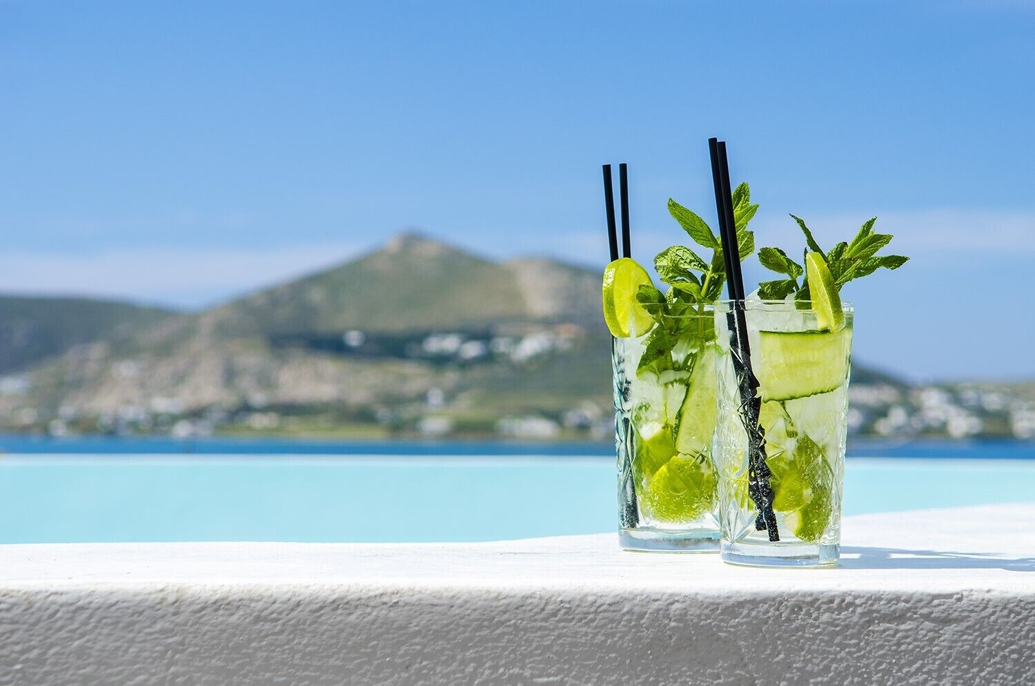 Two glasses of a lime-based drink with mint leaves and black straws, placed on a white surface with a coastal view and mountains in the background.