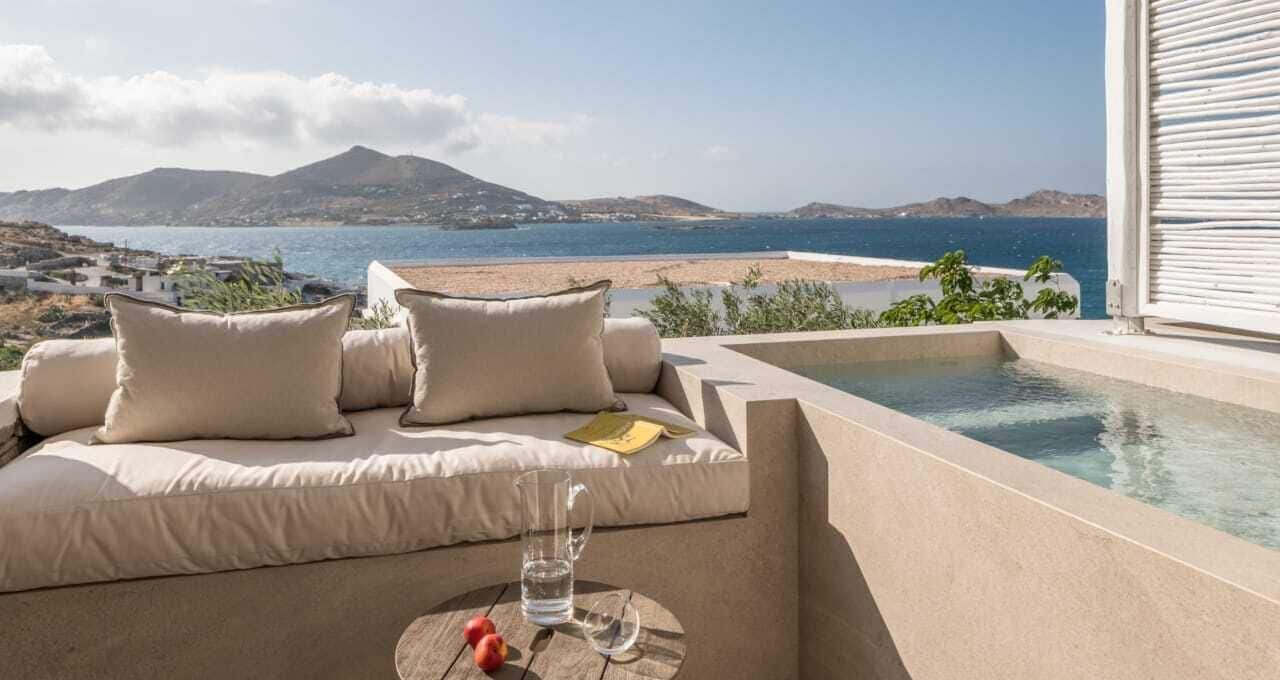 A sunlit outdoor seating area with a pool, overlooking a beach and mountains.
