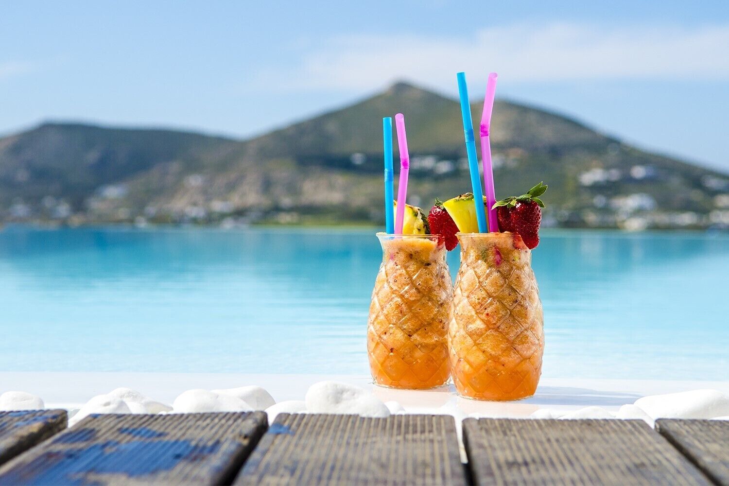 Two glass containers with colorful straws and fruit on a wooden deck by a pool with a mountain in the background.