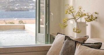 A room with a view of the sea, a glass door, and a vase with flowers on a table.