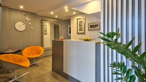A modern reception area with a reception desk, two orange chairs, a clock, framed pictures, and a plant. - Jules Suites (St. Julian's)
