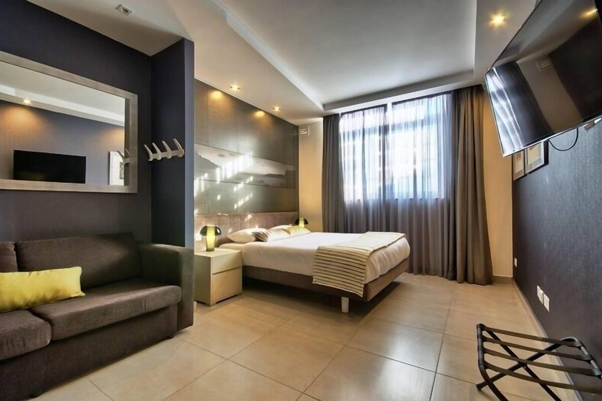 A modern hotel room with a bed, a sofa, a TV, and a large window with curtains.