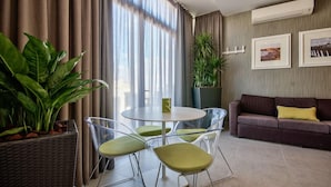 A modern living room with a round glass table, green chairs, a brown sofa with a yellow pillow, and a large window with curtains. - Jules Suites (St. Julian's)
