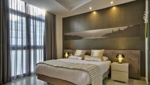 A modern bedroom with a large bed, two bedside tables with lamps, a cityscape painting on the wall, and a view through sheer curtains. - Jules Suites (St. Julian's)