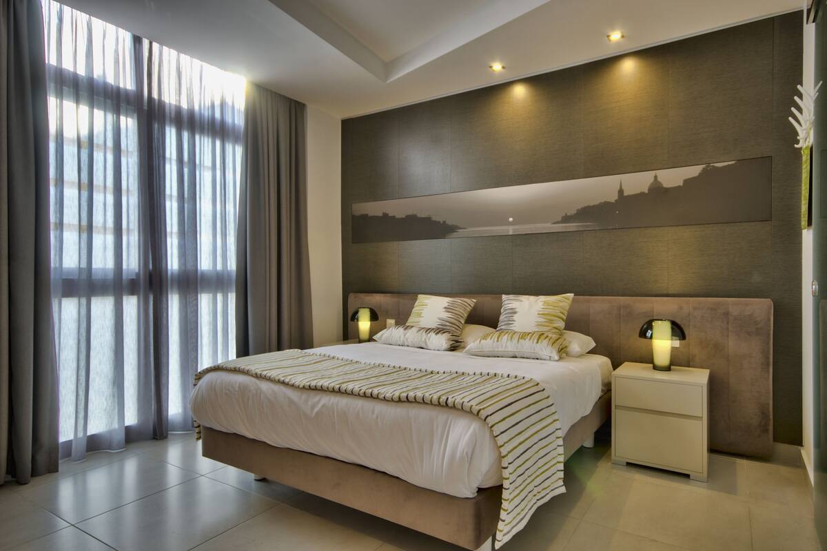 A modern bedroom with a large bed, two bedside tables with lamps, a cityscape painting on the wall, and a view through sheer curtains.