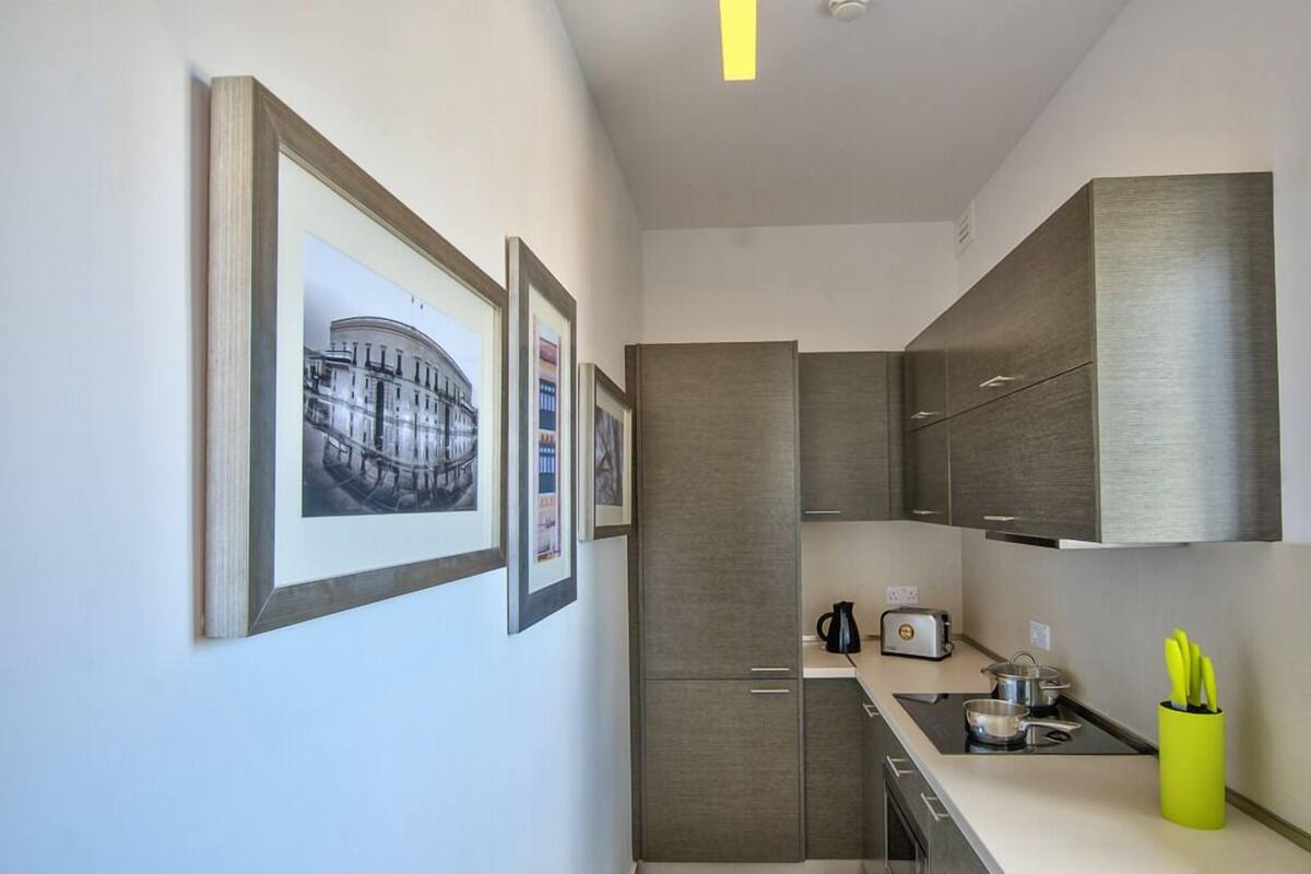 A modern kitchen with grey cabinets, a built-in oven, and a small countertop. There are framed pictures on the wall and a yellow utensil holder on the counter.
