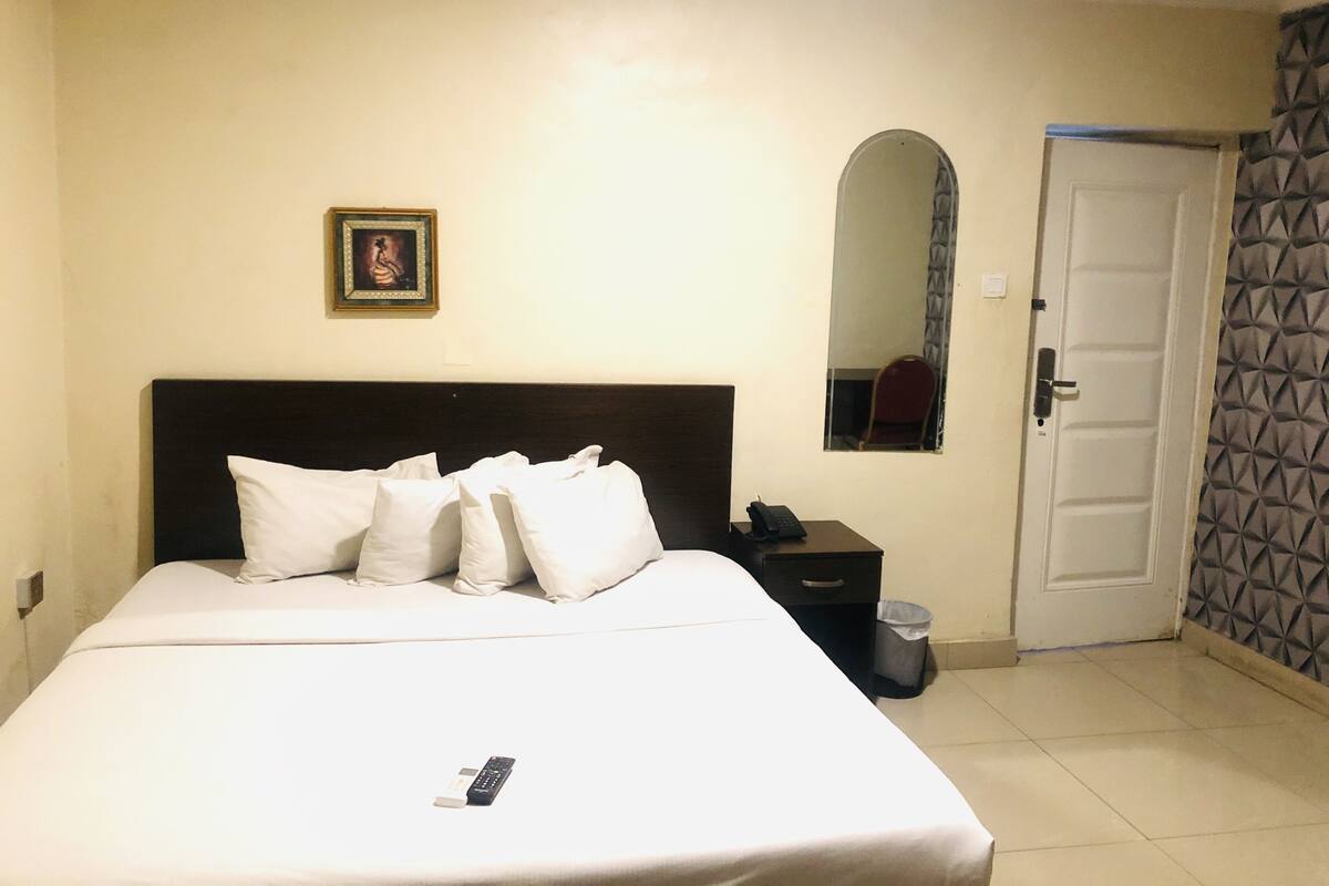 A hotel room with a bed, pillows, a bedside table with a phone, a trash bin, a mirror, and a door.
