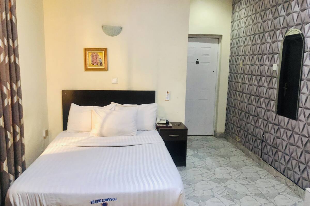 A hotel room with a bed, a nightstand, a wall-mounted light, a door, and a patterned wall.