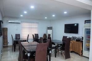 A dining area with a table and chairs, an air conditioning unit, a television, and a refrigerator. - Mayors & Diplomat Suites Opebi (Lagos)