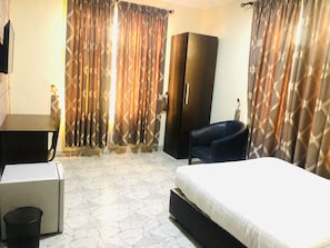 A hotel room with a bed, a desk, a chair, a wardrobe, and curtains. - Mayors & Diplomat Suites Opebi (Lagos)